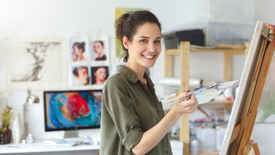Stylish young Caucasian woman with dark hair taking part in class and workshop for artists, feeling happy and excited, standing in studio in front of easel and smiling. Art, learning and education
