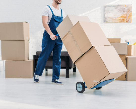 cropped view of mover in uniform transporting cardboard boxes on hand truck in apartment
